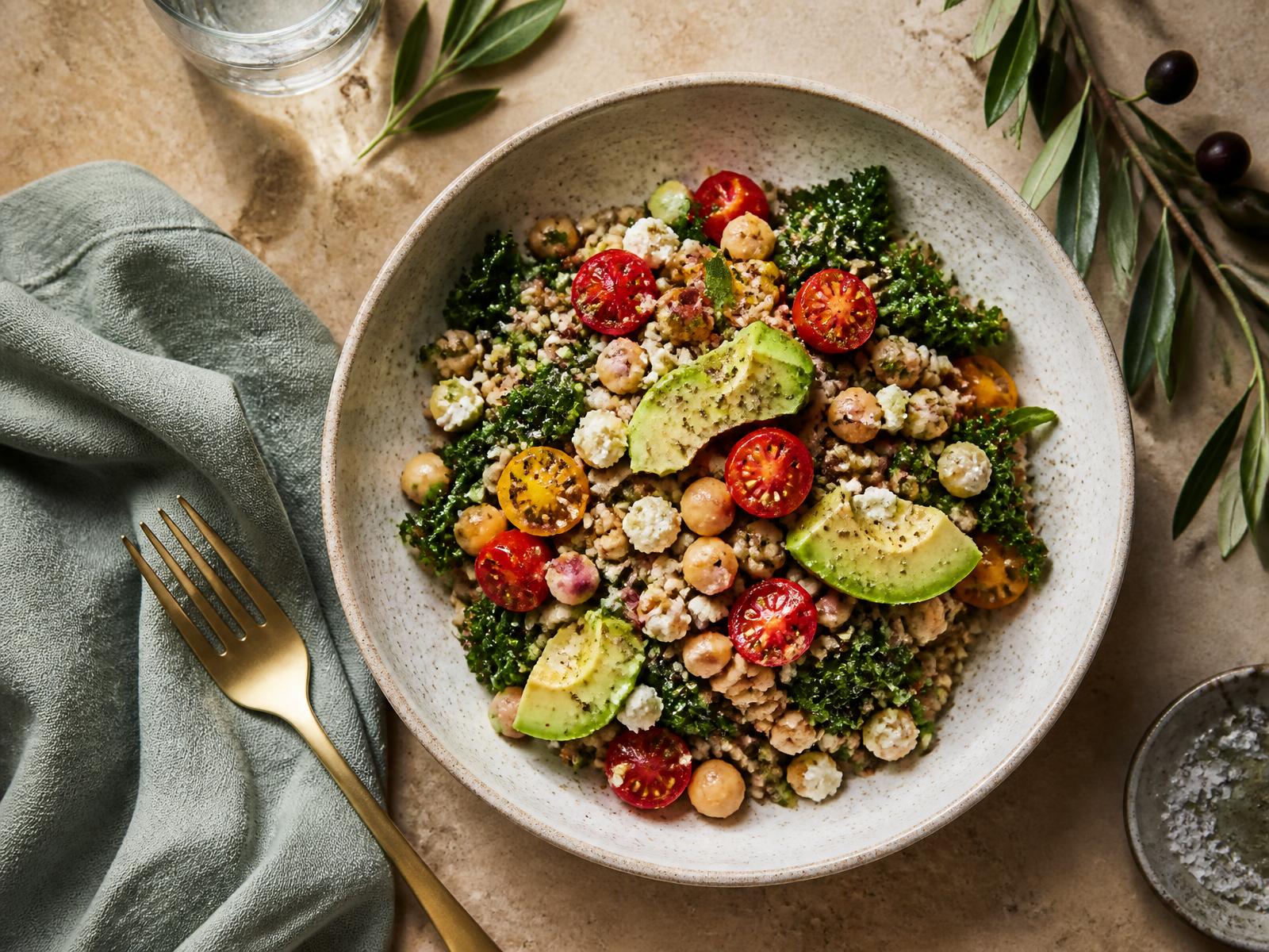A speckled ceramic bowl of grains, chickpeas, tomatoes, kale and avocado on a warm stone table